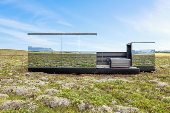 A modern, minimalist structure with a glass enclosure, a wooden platform, and a metal bench in a grassy field with distant mountains. - Heathland Lodge (Selfoss)