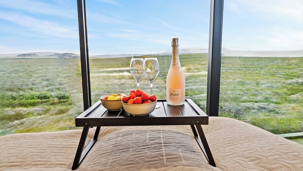 A bed with a view of a grassy landscape through large windows, a bottle of champagne, two glasses, and a bowl of strawberries. - Heathland Lodge (Selfoss)