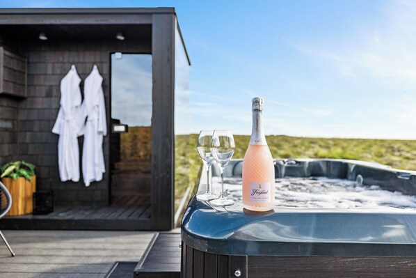 A hot tub with a bottle of champagne and two glasses, a wooden deck, and a small building with white robes hanging on the wall. - Heathland Lodge (Selfoss)