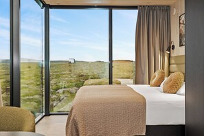A modern hotel room with a large window, a bed with patterned bedding, and a view of a grassy landscape. - Heathland Lodge (Selfoss)