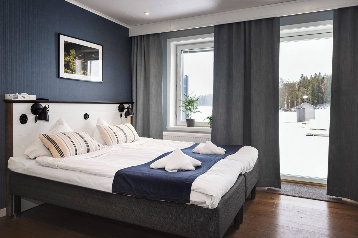 Cama arrumada com almofadas e um cobertor, mesa de cabeceira com abajur, janela com cortinas e um quadro na parede.