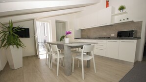 Cocina moderna con gabinetes blancos, encimera gris y microondas integrado. Hay una mesa de comedor con sillas y una planta en maceta.