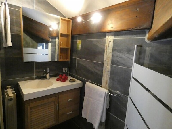 Un baño moderno con un espejo grande, paredes cubiertas de baldosas oscuras, un lavabo blanco, armarios de madera y un perchero para toallas.