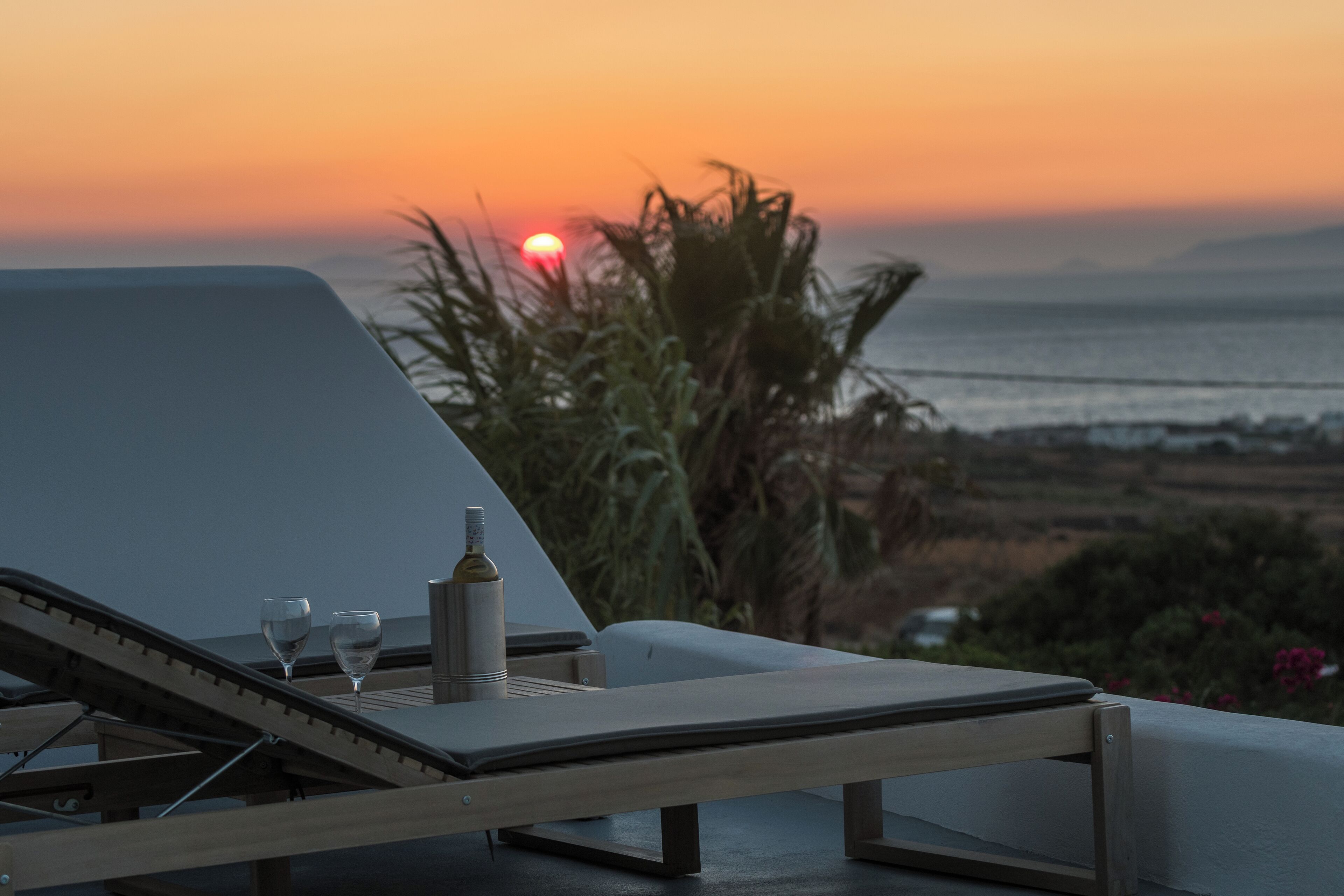 Un área de descanso iluminada por el sol con vista a la puesta de sol sobre el mar, una botella y dos vasos sobre una mesa.