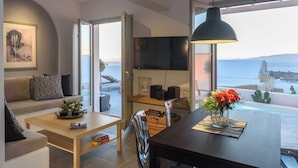 Una sala moderna con sofá, mesa de centro y comedor con mesa y sillas. Hay un televisor montado en la pared y un ventanal con vistas al mar.