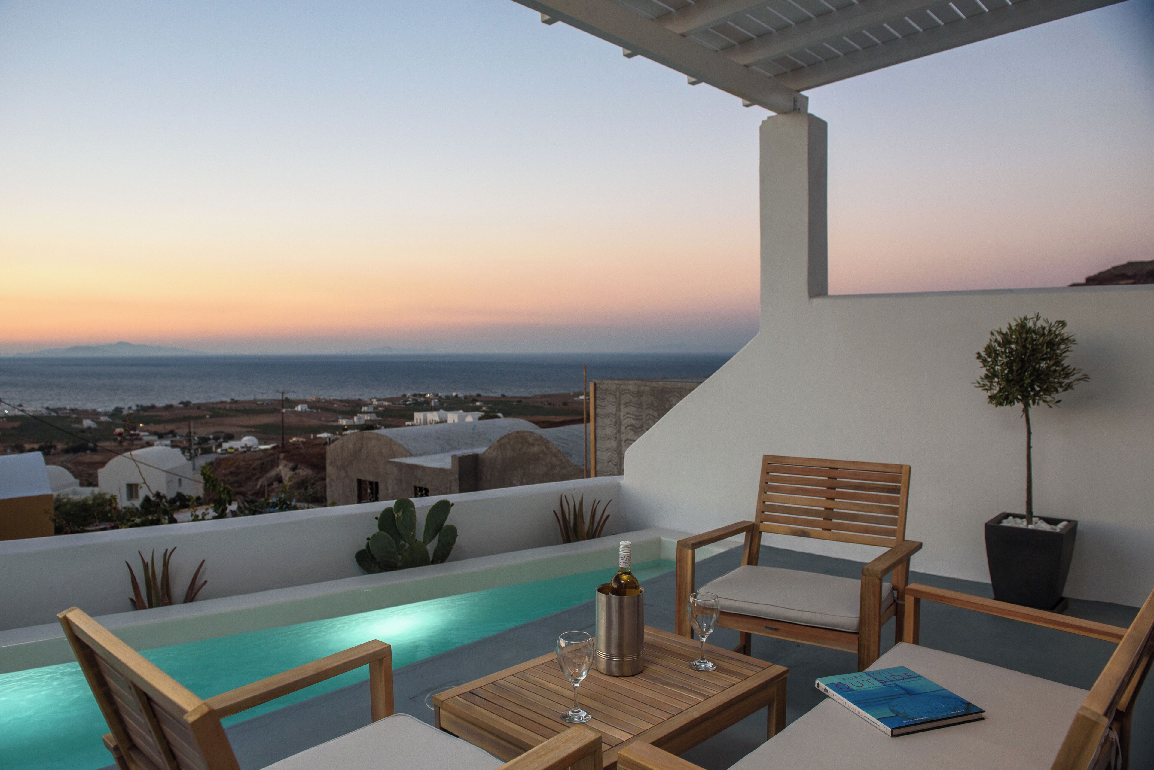 A terrace with a pool, wooden furniture, and a view of the sea and distant buildings.