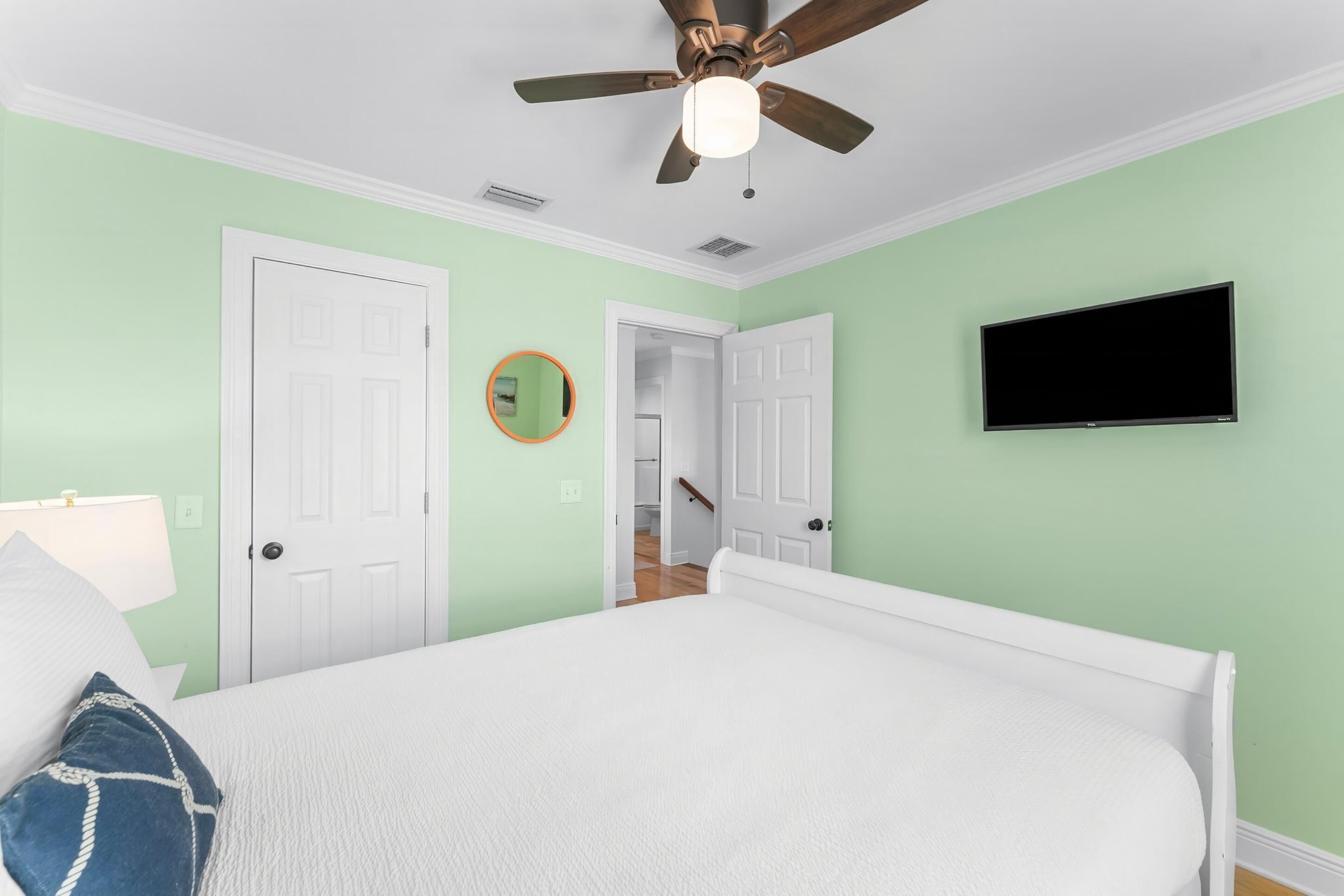 A bedroom with a bed, a ceiling fan, a television, and a door leading to another room.