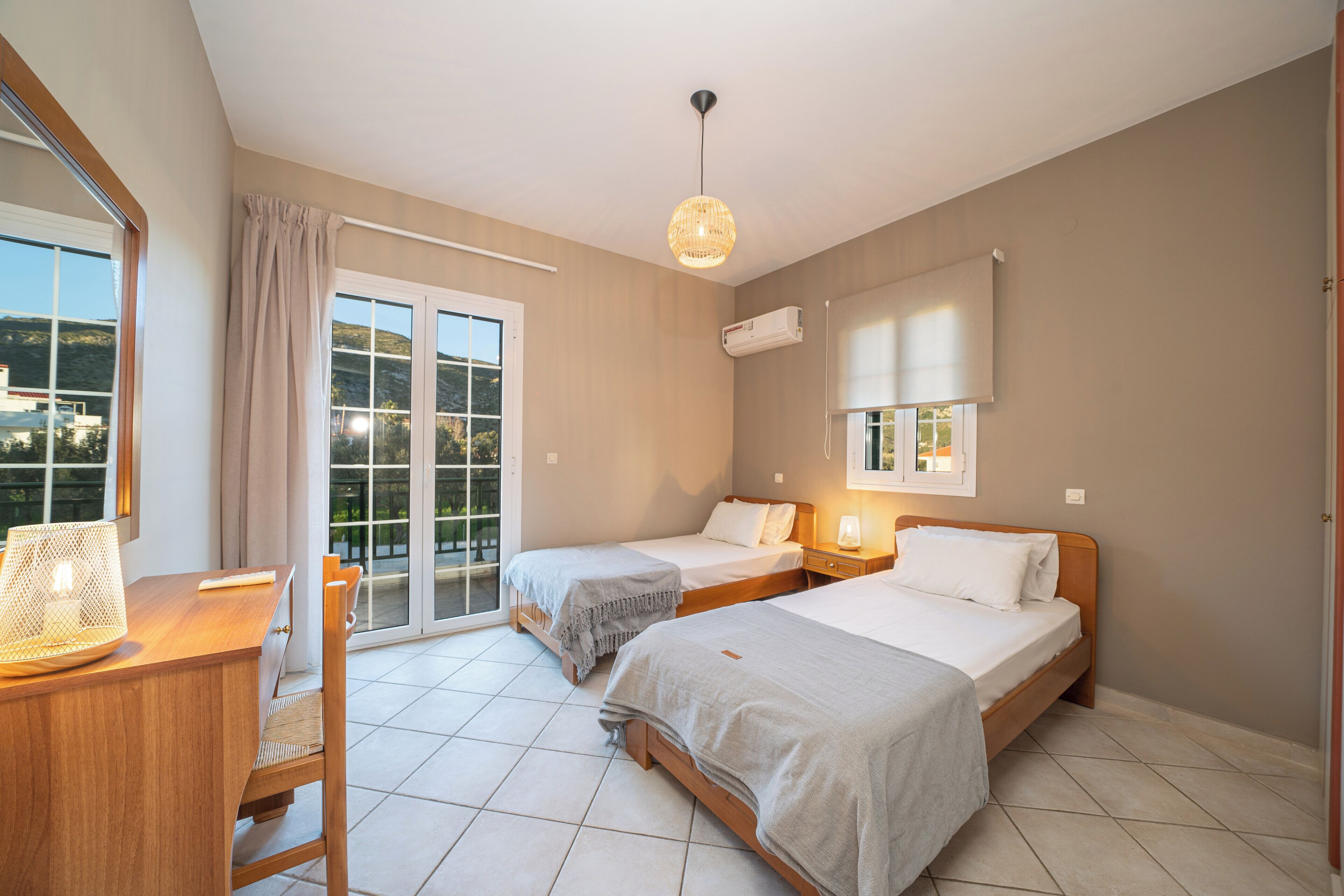 A hotel room with two beds, a desk, a chair, a balcony with a view, and a hanging light fixture.