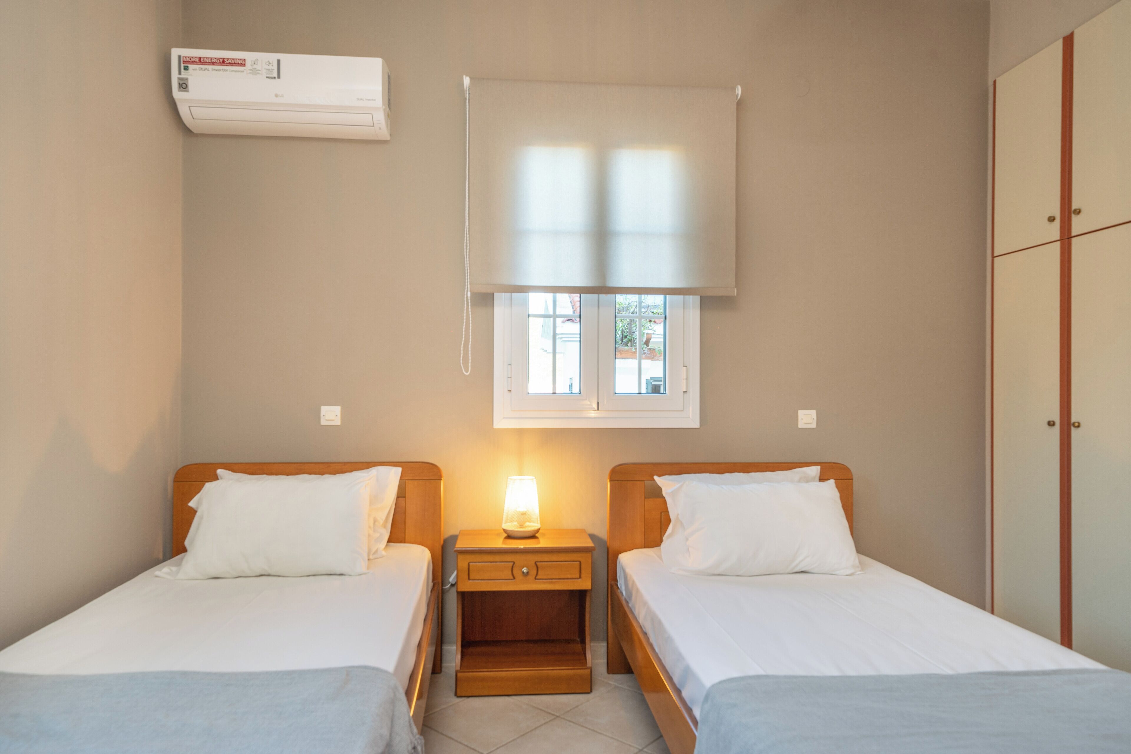 A room with two beds, a nightstand with a lamp, a window with blinds, and an air conditioning unit on the wall.