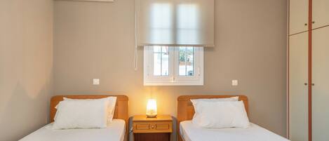 A room with two beds, a nightstand with a lamp, a window with blinds, and an air conditioning unit on the wall.