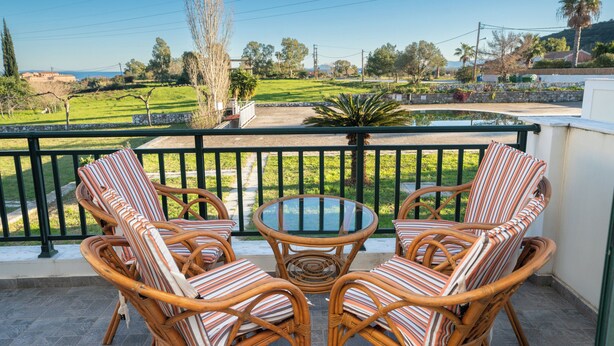 A balcony with wicker furniture and a glass table, overlooking a green landscape with trees and a distant view of hills. - Captain's Villas (Kefalonia)
