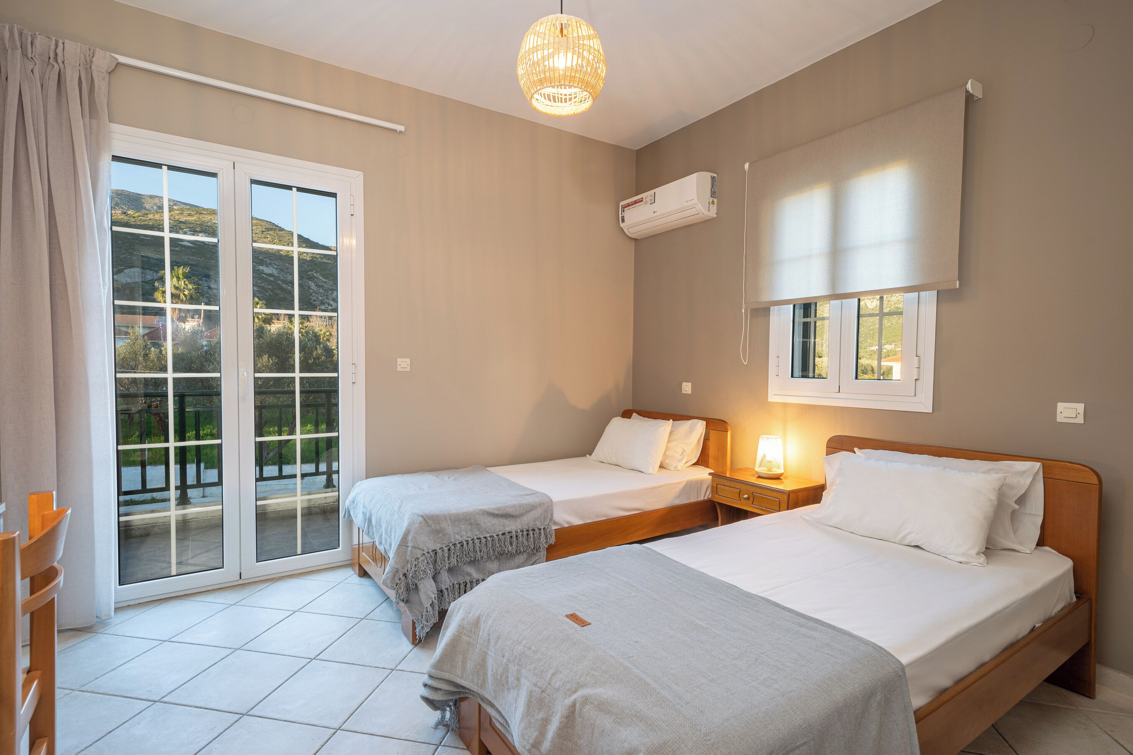 A hotel room with two single beds, a sliding glass door to the outside, an air conditioning unit, and a bedside table with a lamp.