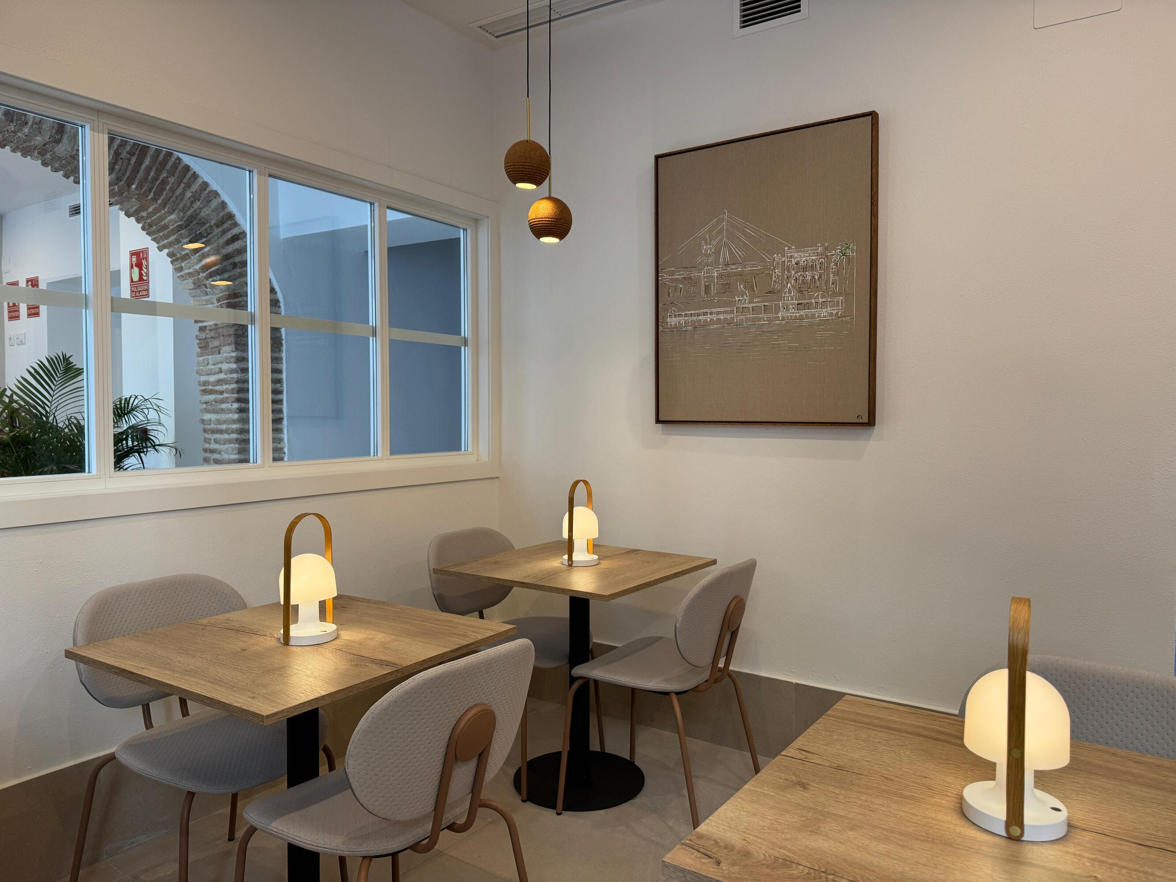 Un'area pranzo moderna con tavoli e sedie in legno, lampade a sospensione e un'ampia finestra che si affaccia su un edificio e palme.