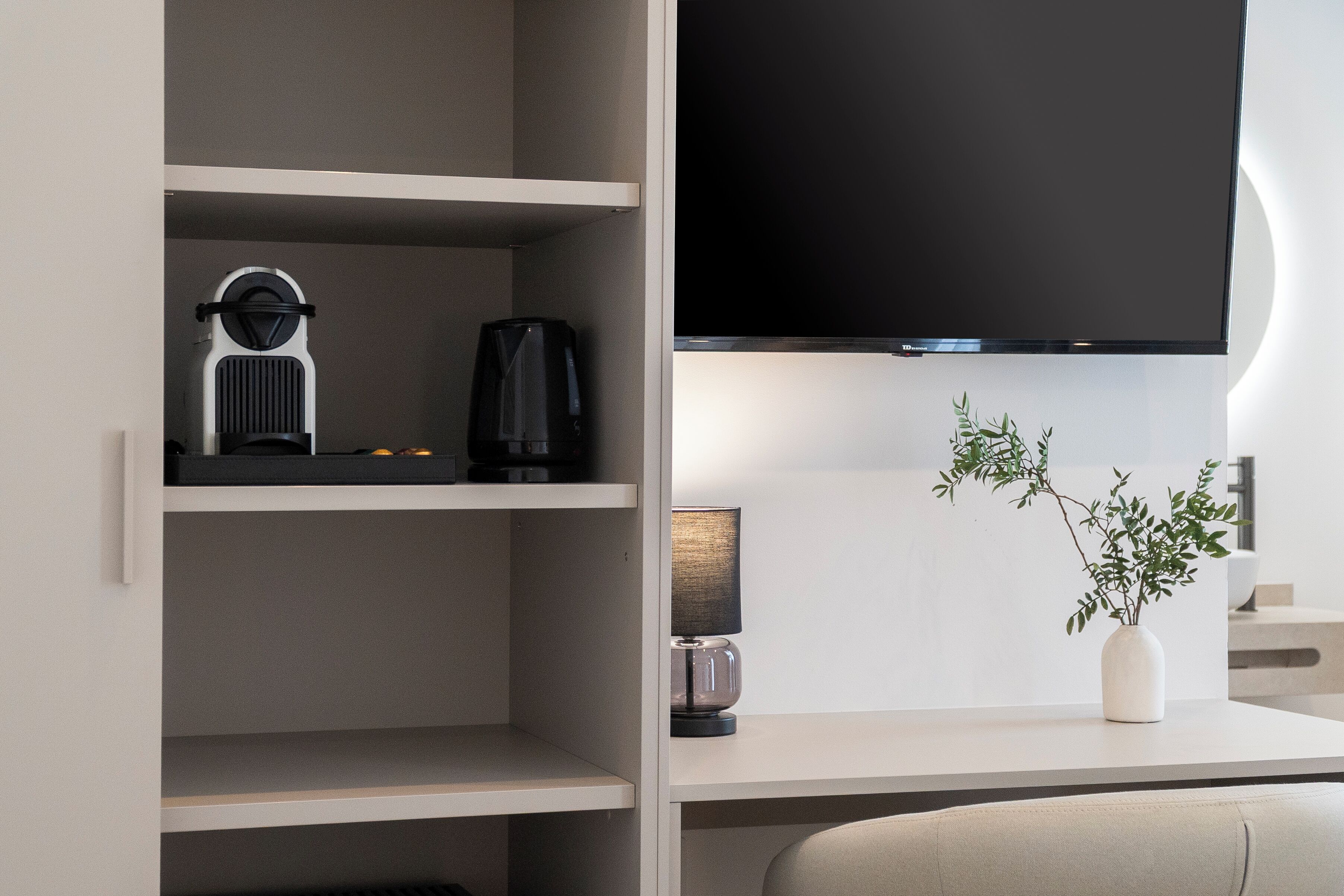 A modern living room with a flat-screen TV mounted on the wall, a coffee maker, and a small table with a lamp and a plant.