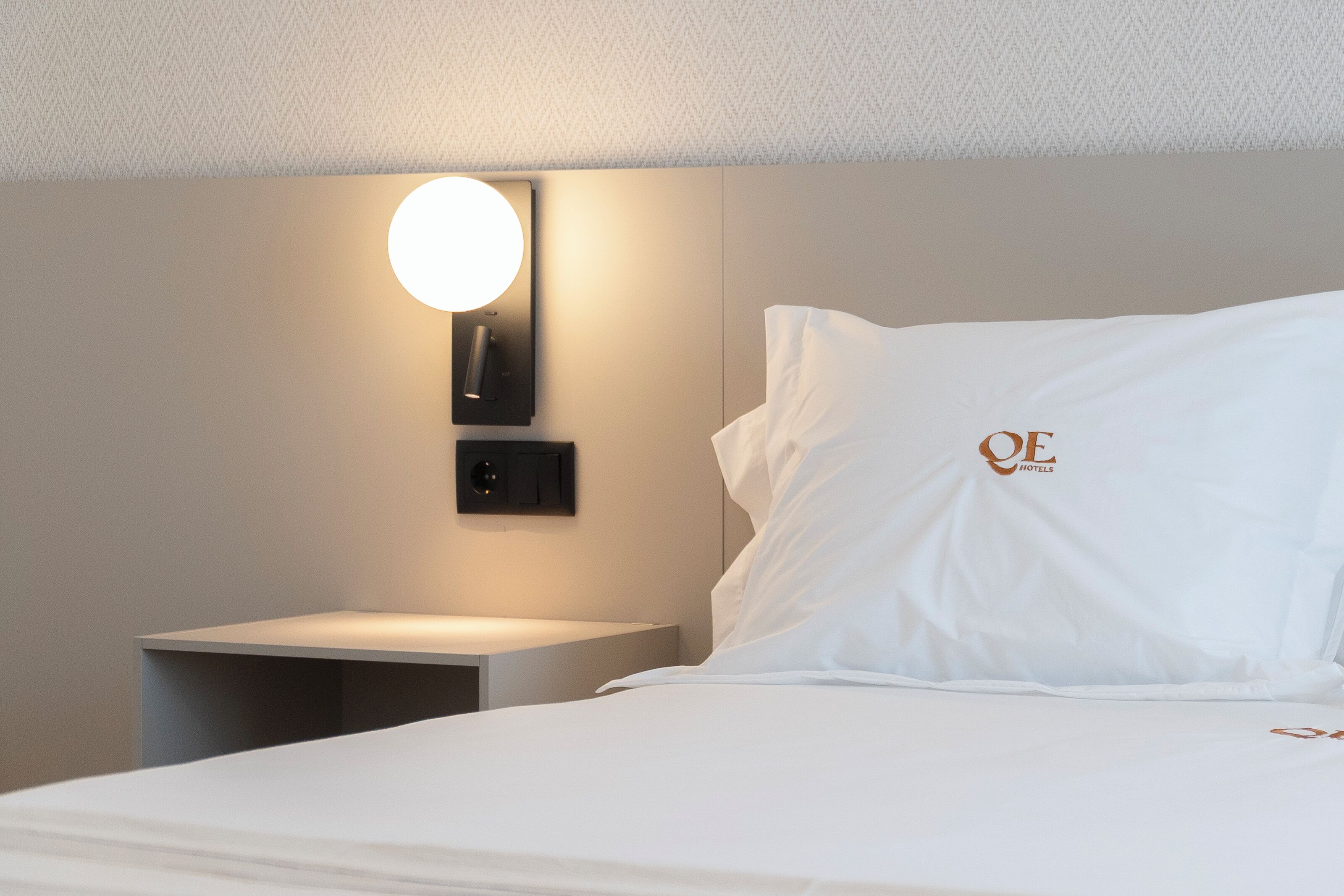 A hotel room with a neatly made bed, a bedside table, a wall-mounted light, and a pillow with a logo.