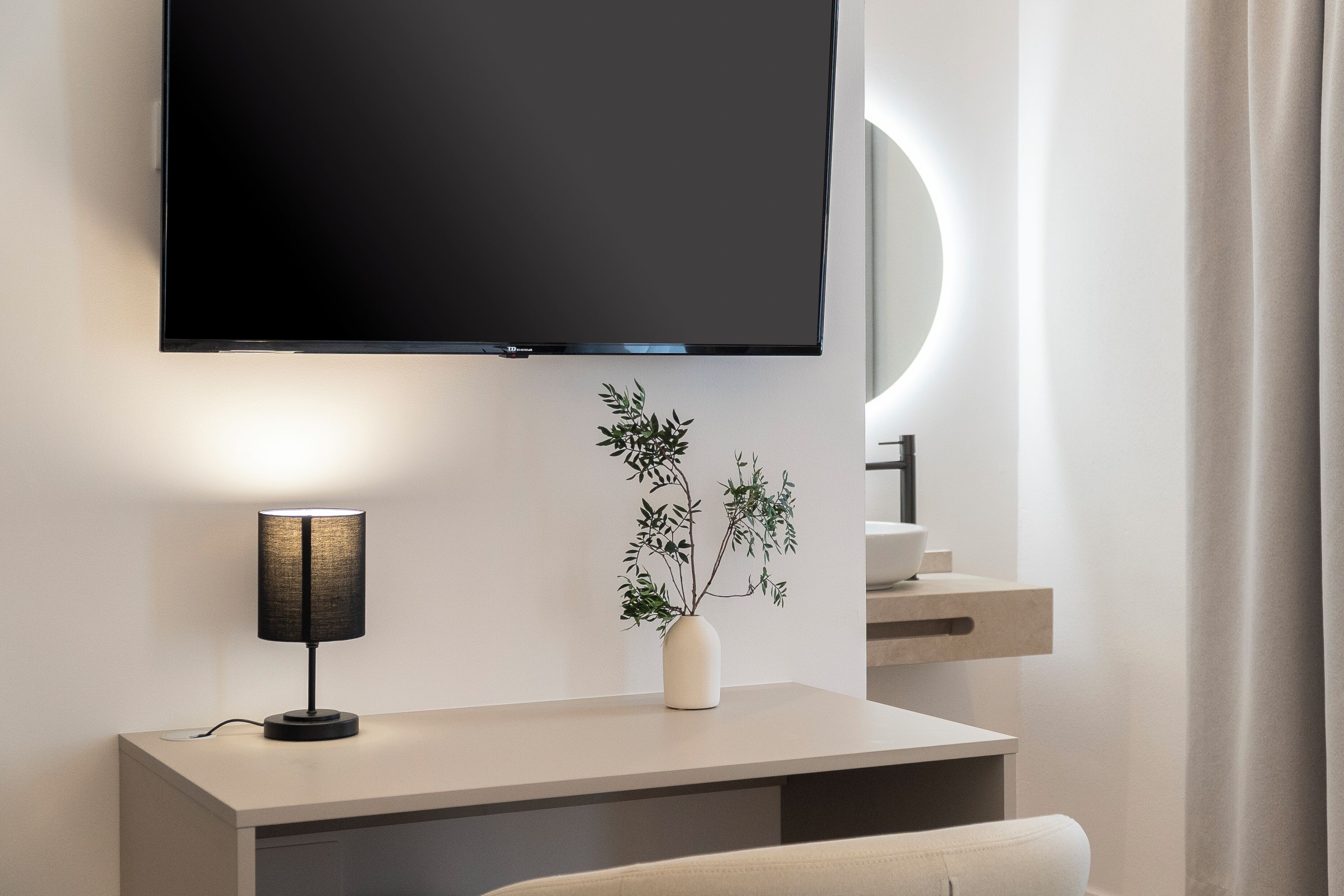 Ein modernes Hotelzimmer mit flachbildfernseher an der Wand, einem Nachttisch mit Lampe, einer Pflanze im Blumenvasen und minimalistischem Design.