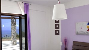 Ein Schlafzimmer mit Bett, Nachttisch, Lampe, einem Gemälde und Meerblick durch eine Glastür.