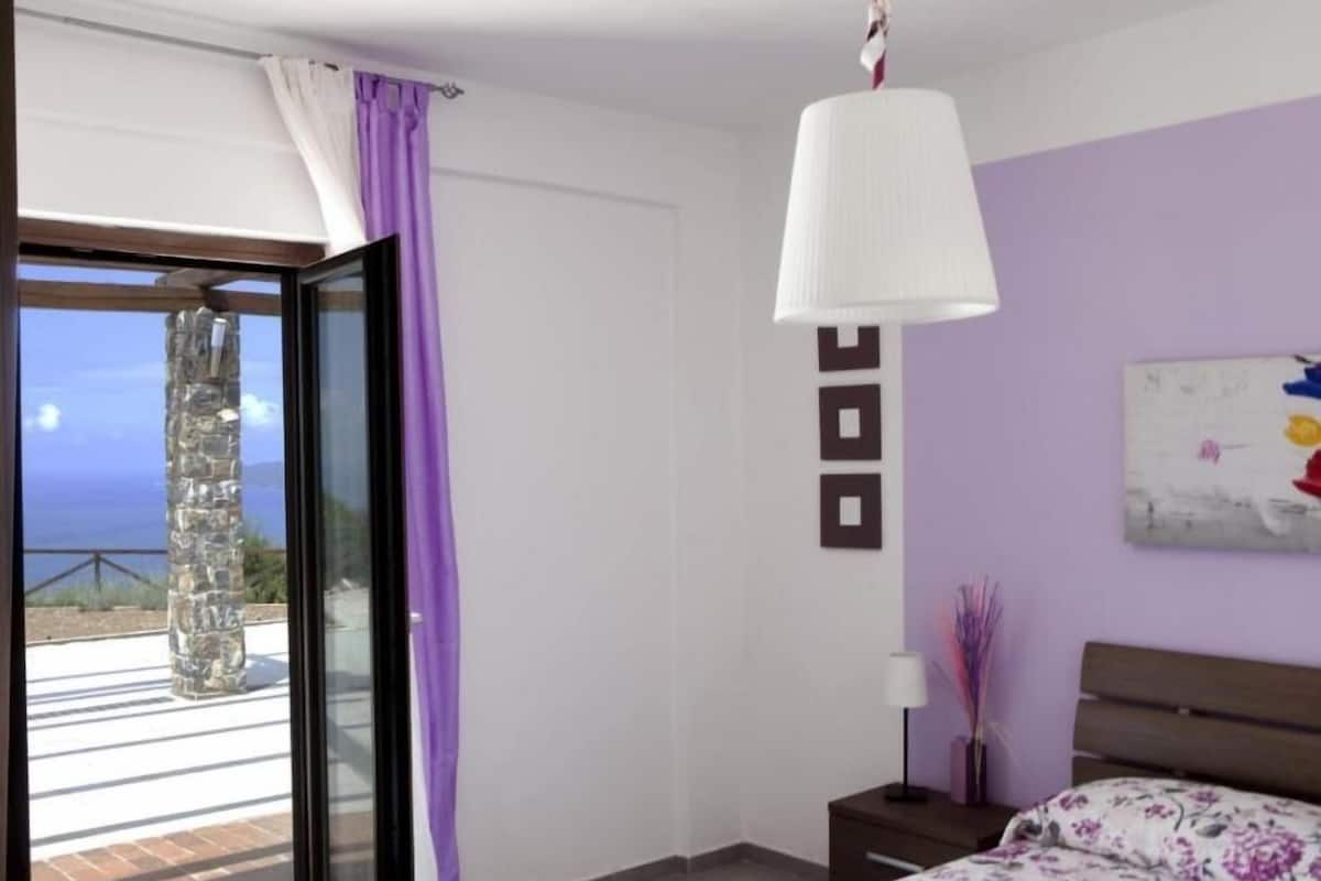 Une chambre à coucher avec un lit, une table de chevet, une lampe, un tableau et une vue sur la mer, visible à travers une porte vitrée.