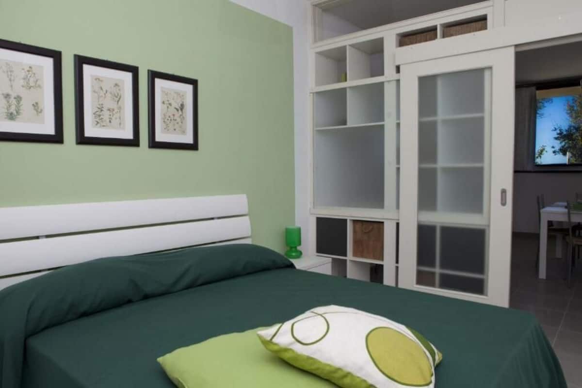 Une chambre à coucher avec un lit, une literie verte, un oreiller vert, une tête de lit blanche et un mur orné d’illustrations botaniques encadrées.