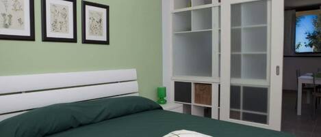 A bedroom with a bed, green bedding, a green pillow, a white headboard, and a wall with framed botanical illustrations.
