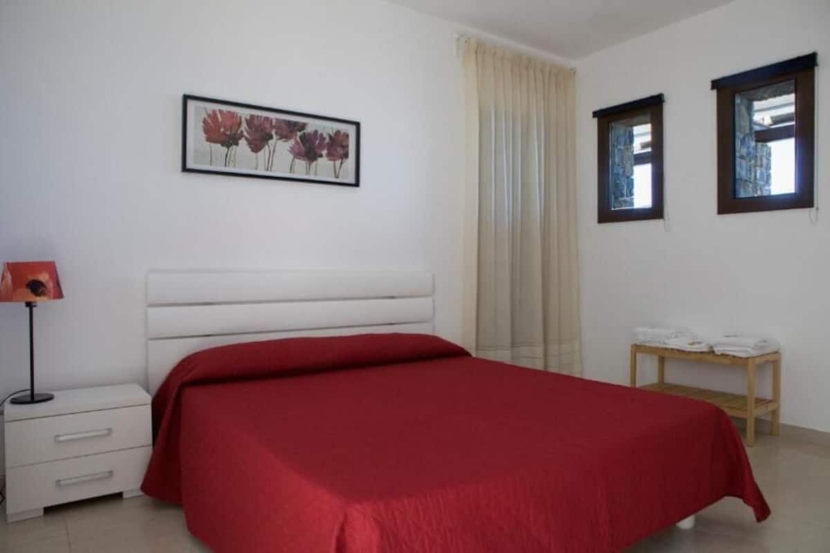 Une chambre à coucher avec un grand lit, une table de chevet avec une lampe, un tableau encadré au mur et une fenêtre avec des rideaux.