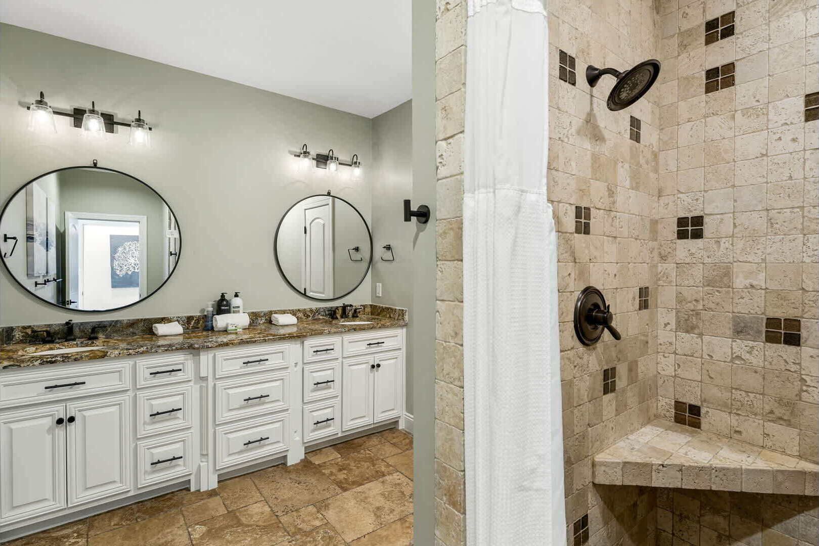 Un baño con encimera de granito, muebles de color blanco, dos espejos redondos y una ducha con pared de piedra.