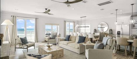 A spacious living room with a large sofa, armchairs, and a coffee table. There are ceiling fans, a kitchen area with a dining table, and large windows offering a view of the ocean.