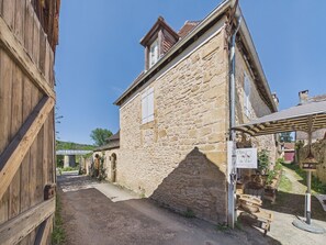 A stone building with a sloped roof and a small window, a dirt road leading to a bridge, and a sign indicating a local artisan market.