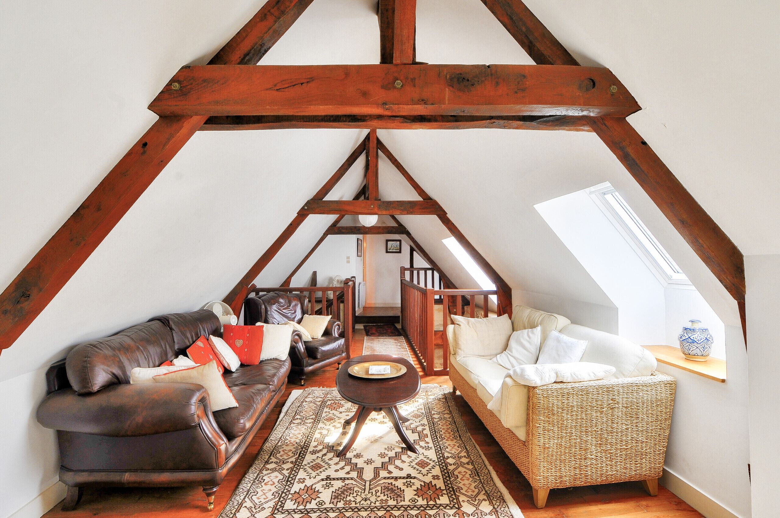 A loft-style room with a high ceiling, wooden beams, a brown leather sofa, a wicker chair, and a patterned rug.