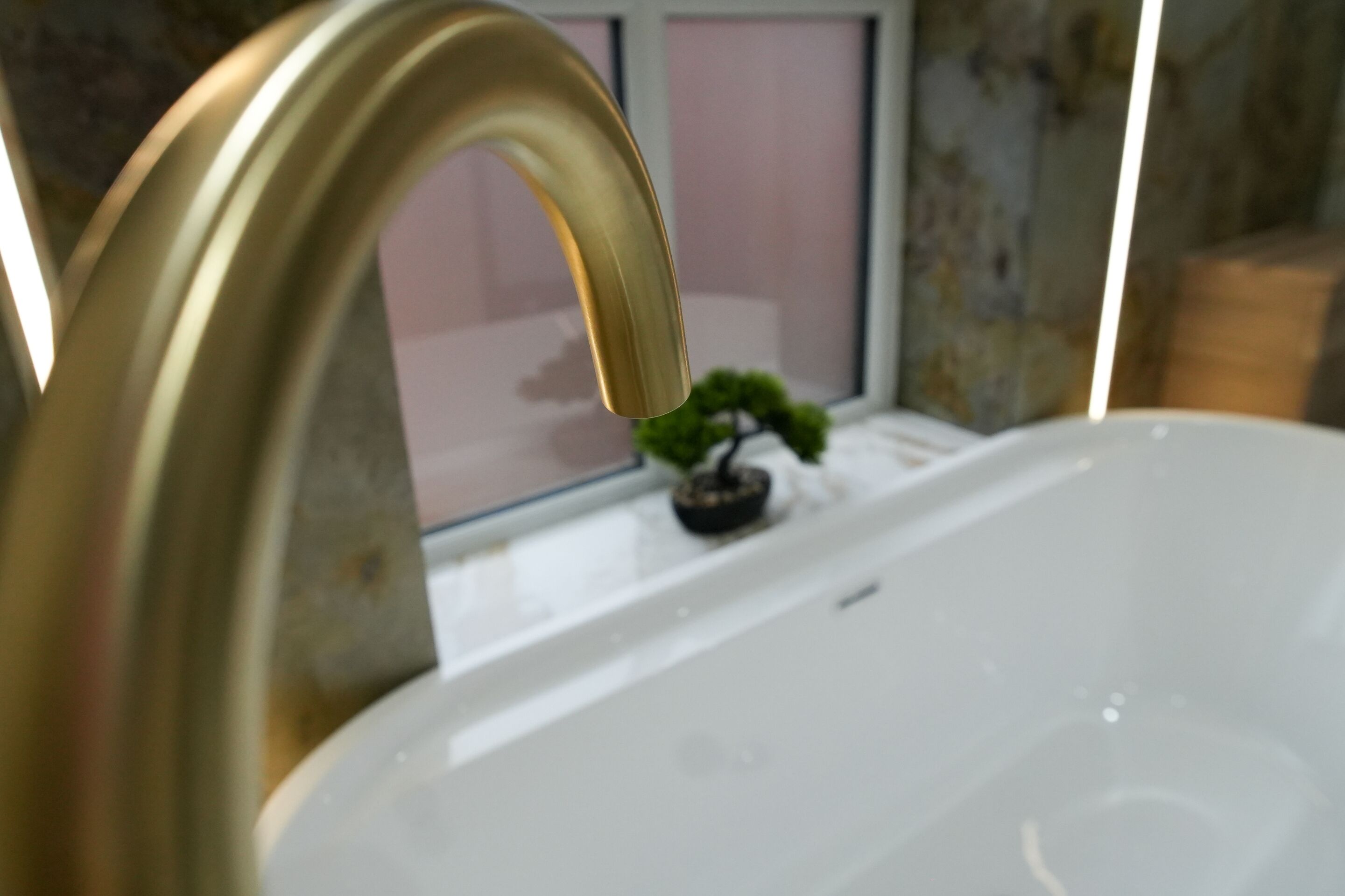 A bathroom with a gold faucet, a white sink, and a small potted plant on the windowsill.
