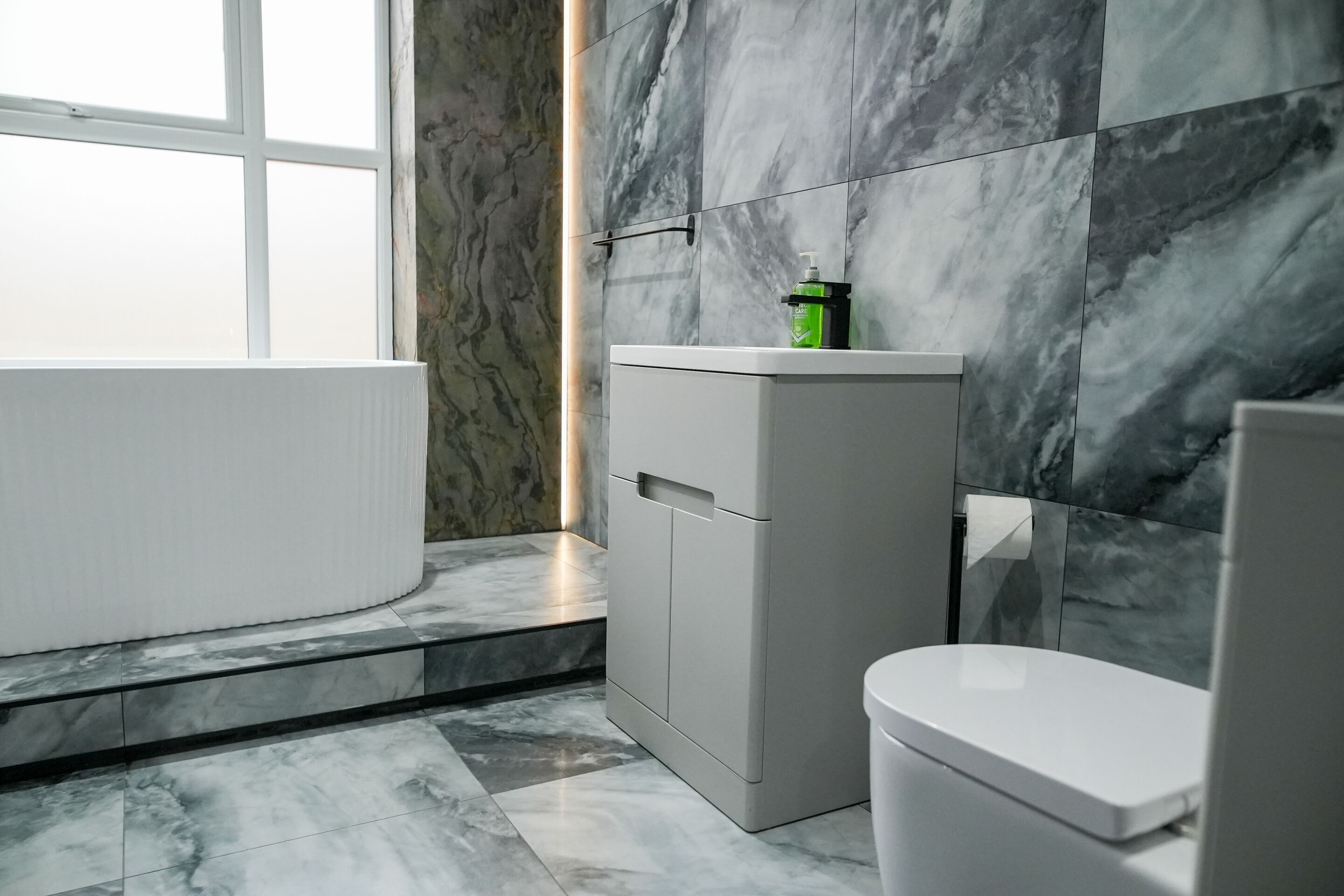Een moderne badkamer met een marmeren vloer, een witte badkuip, een toilet en een kastje met een groene fles erop.