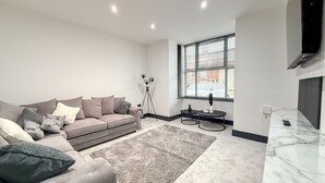 A modern living room with a grey sofa, a coffee table, and a large window with blinds. - Santi House (Boston)
