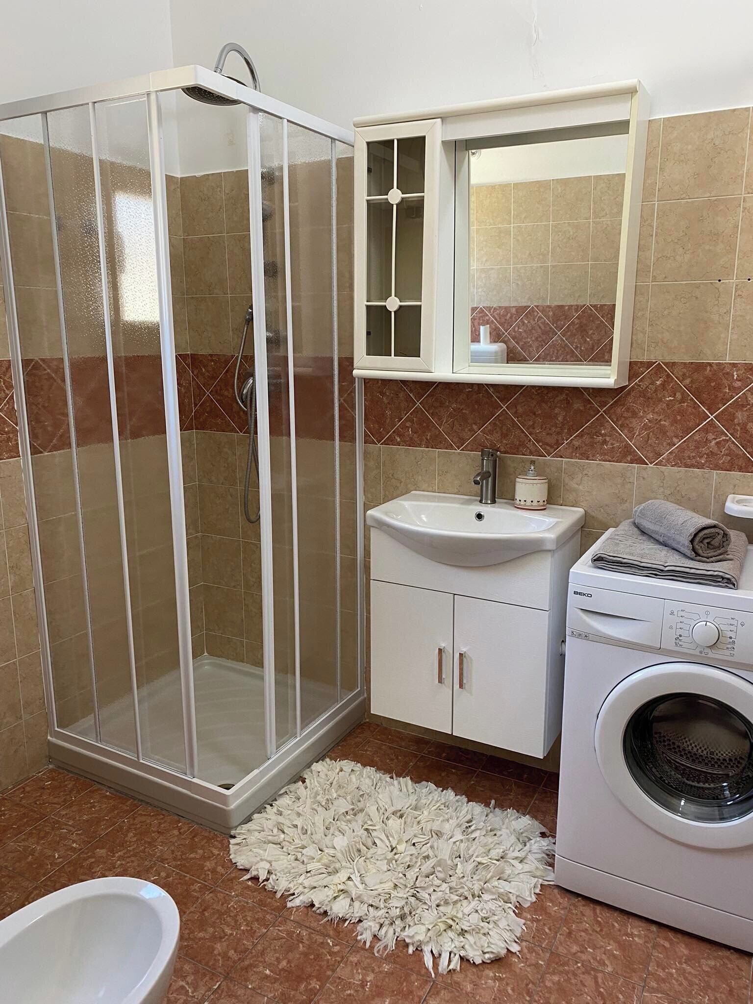 A bathroom with a glass-enclosed shower, a sink, a mirror, a washing machine, and a toilet.