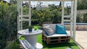 Een overdekt terras met een houten bank en lounge fauteuils, omgeven door groen en uitzicht op het landschap.