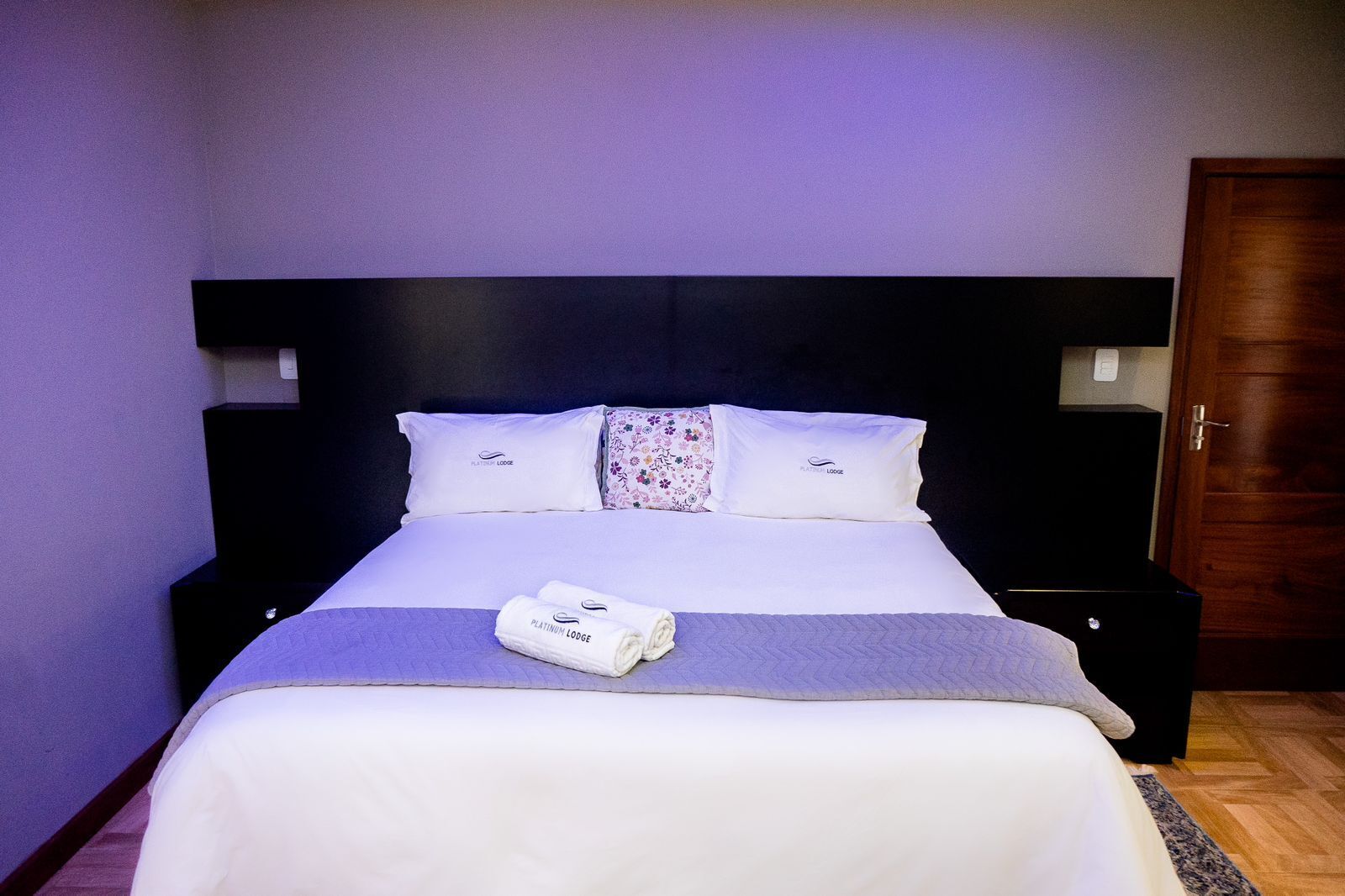 A neatly made bed with white linens and a purple blanket, two pillows, a floral pillow, and a folded towel with the hotel logo.