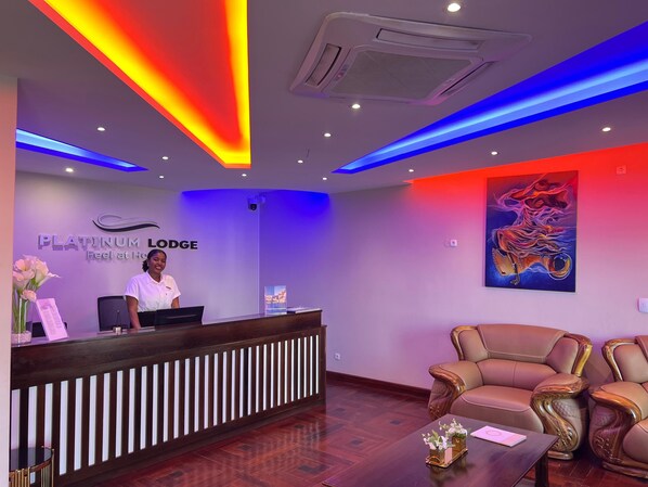 A reception area with a receptionist desk, a seating area with two chairs, and a wall-mounted artwork. - Platinum Lodge (Matola)
