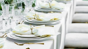 A long table set with white plates, gold cutlery, and clear glassware, arranged in a row with a green backdrop. - Platinum Lodge (Matola)
