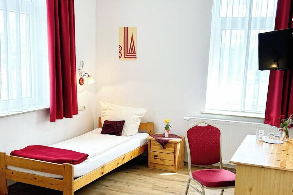 A compact room with a single bed, a desk, and a chair. There are two windows with curtains, a wall-mounted light, and a small table with a vase.