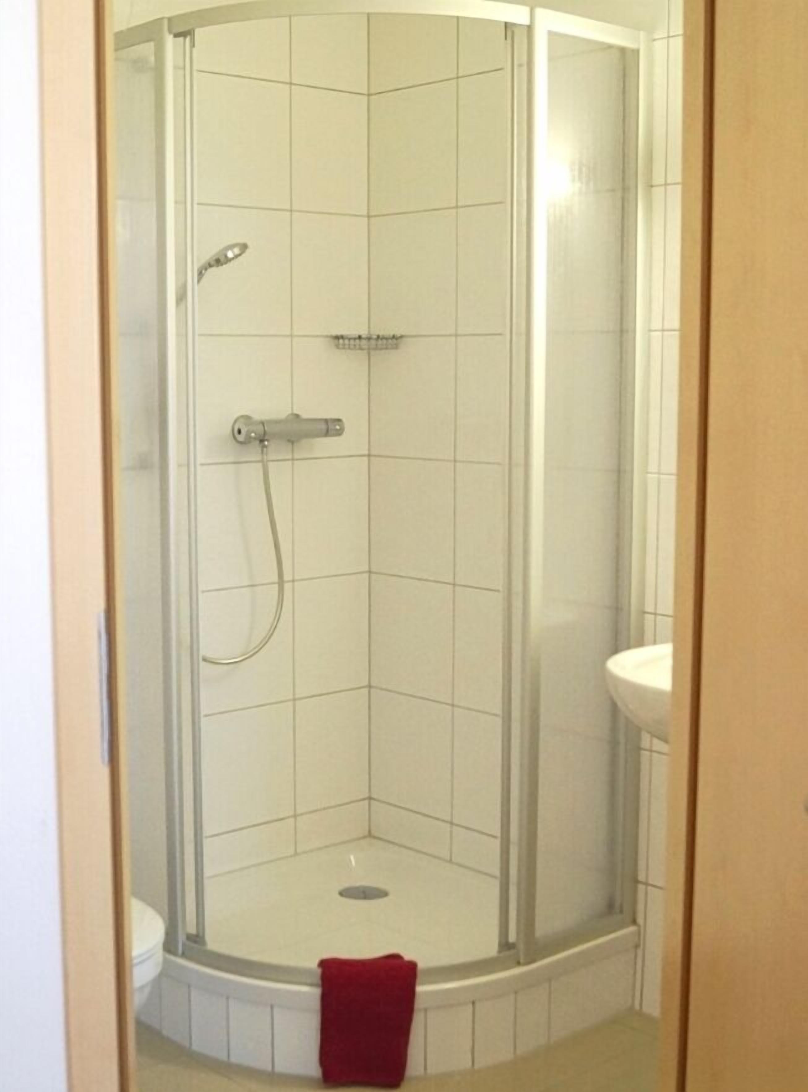A corner shower with a glass door, a white tiled floor, and a red towel hanging on the door.
