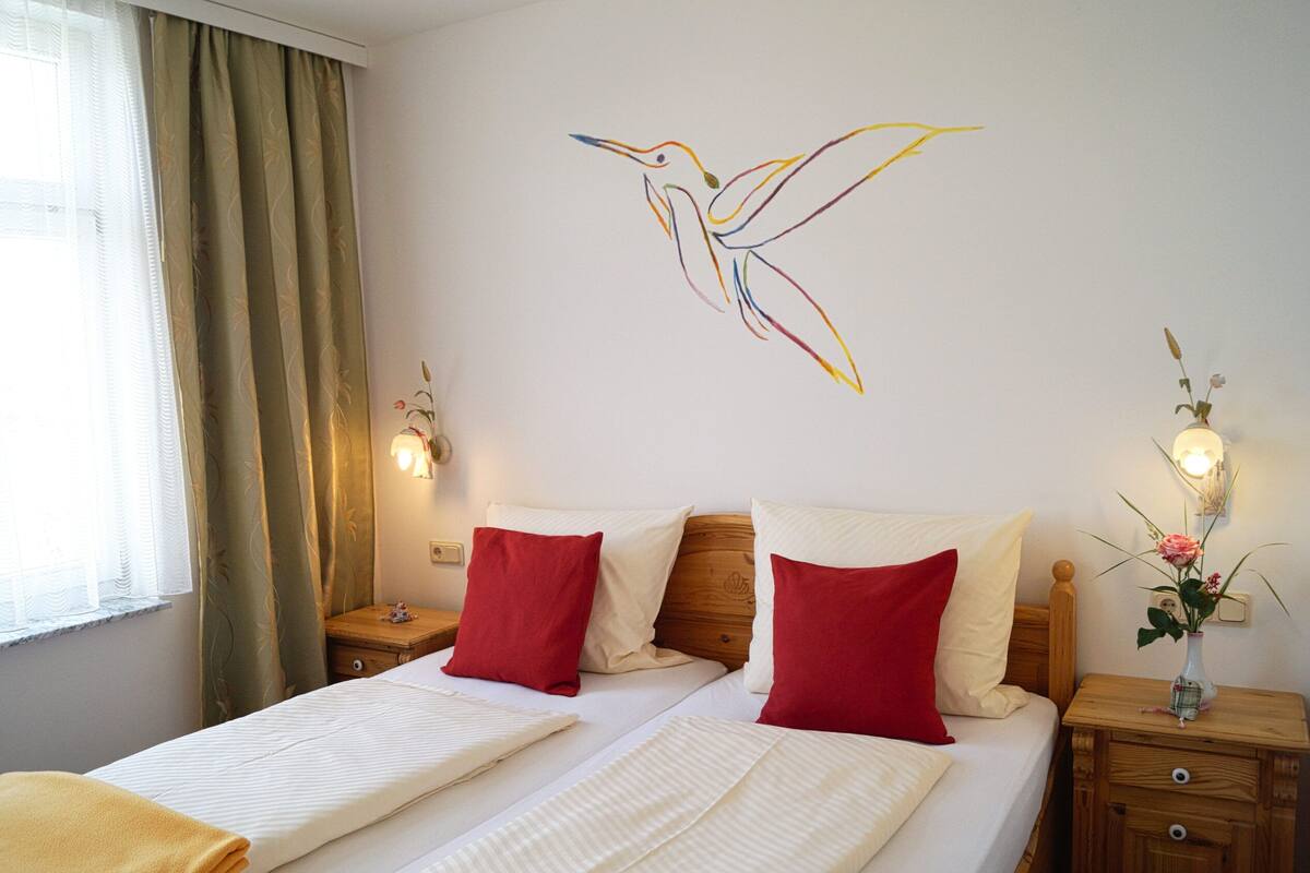 A double bed with red and white pillows, a wooden headboard, and a nightstand with a vase of flowers.
