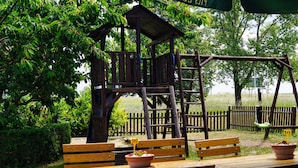 A wooden play structure with a slide and climbing elements, surrounded by benches and picnic tables in a grassy area. - Fürstenhof Landgasthaus und Hotel (Erfurt)
