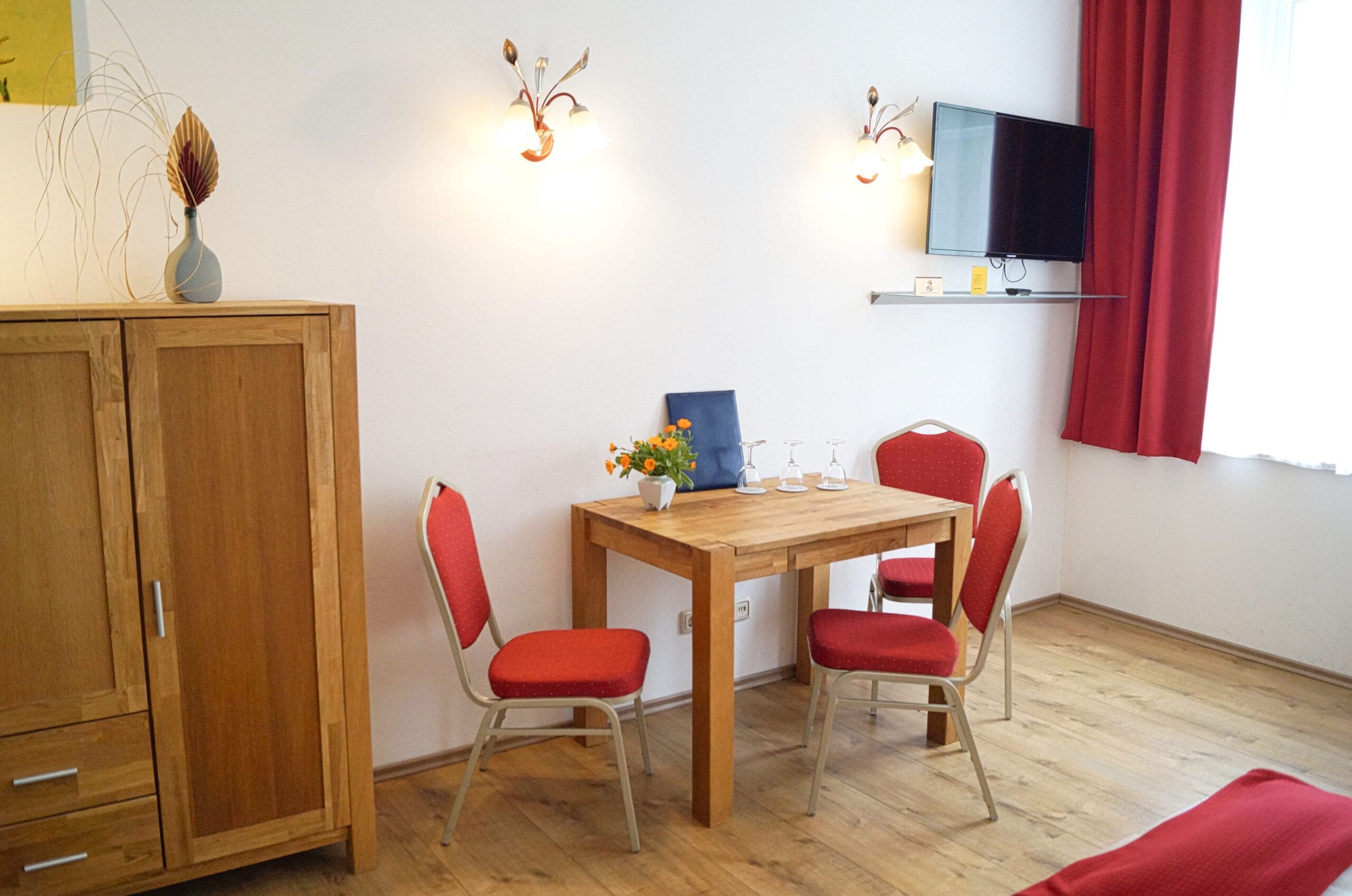 A room with a wooden cabinet, a wooden table with four red chairs, a blue painting, and a television on a shelf.