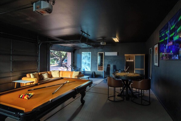 A pool table with a billiard cue and balls, a sofa with cushions, a dining table with chairs, and a large screen displaying an image. - 5 Bed, 3 Bath Uptown Sedona-Game Room-Sauna-Cold Plunge-Jacuzzi-Gym (Sedona)