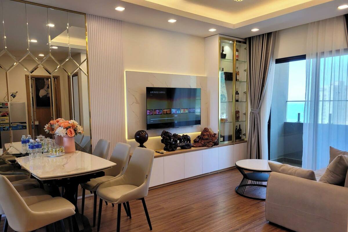 A modern living room with a dining area, a sofa, a TV mounted on the wall, and a glass cabinet with bottles.
