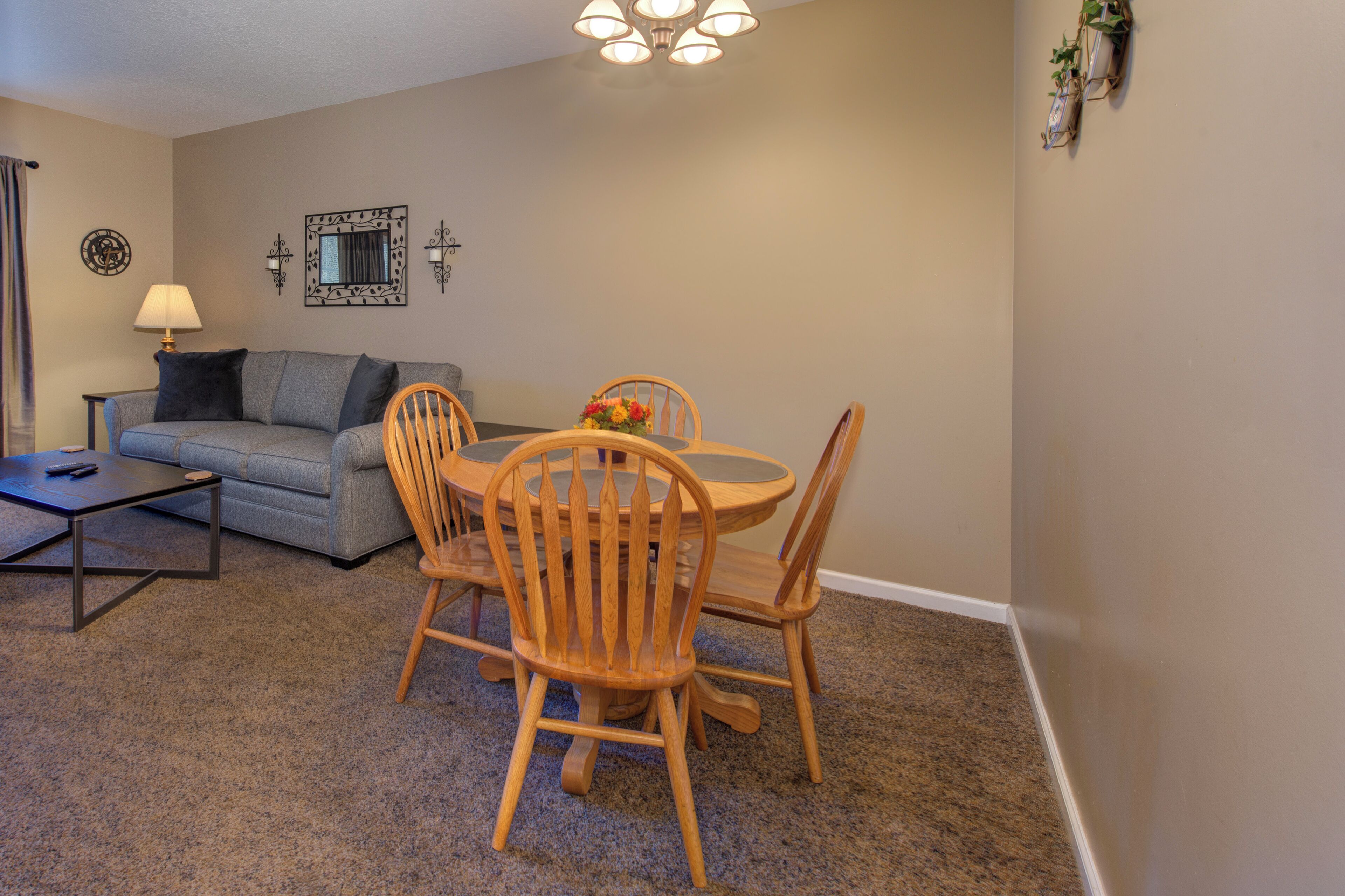 A living room with a grey sofa, a coffee table, a dining table with four wooden chairs, a lamp, and wall-mounted decorations.
