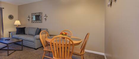 A living room with a grey sofa, a coffee table, a dining table with four wooden chairs, a lamp, and wall-mounted decorations.