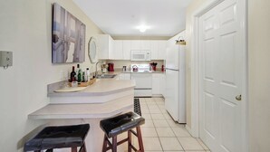 A modern kitchen with white cabinets, a white refrigerator, a microwave, and a stove. There are two black bar stools and a small countertop with bottles and a kettle. - Vanderbilt Villas (Naples)