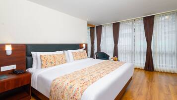 Habitación de hotel con una cama grande, piso de madera, una mesita de noche con teléfono y cortinas en las ventanas.