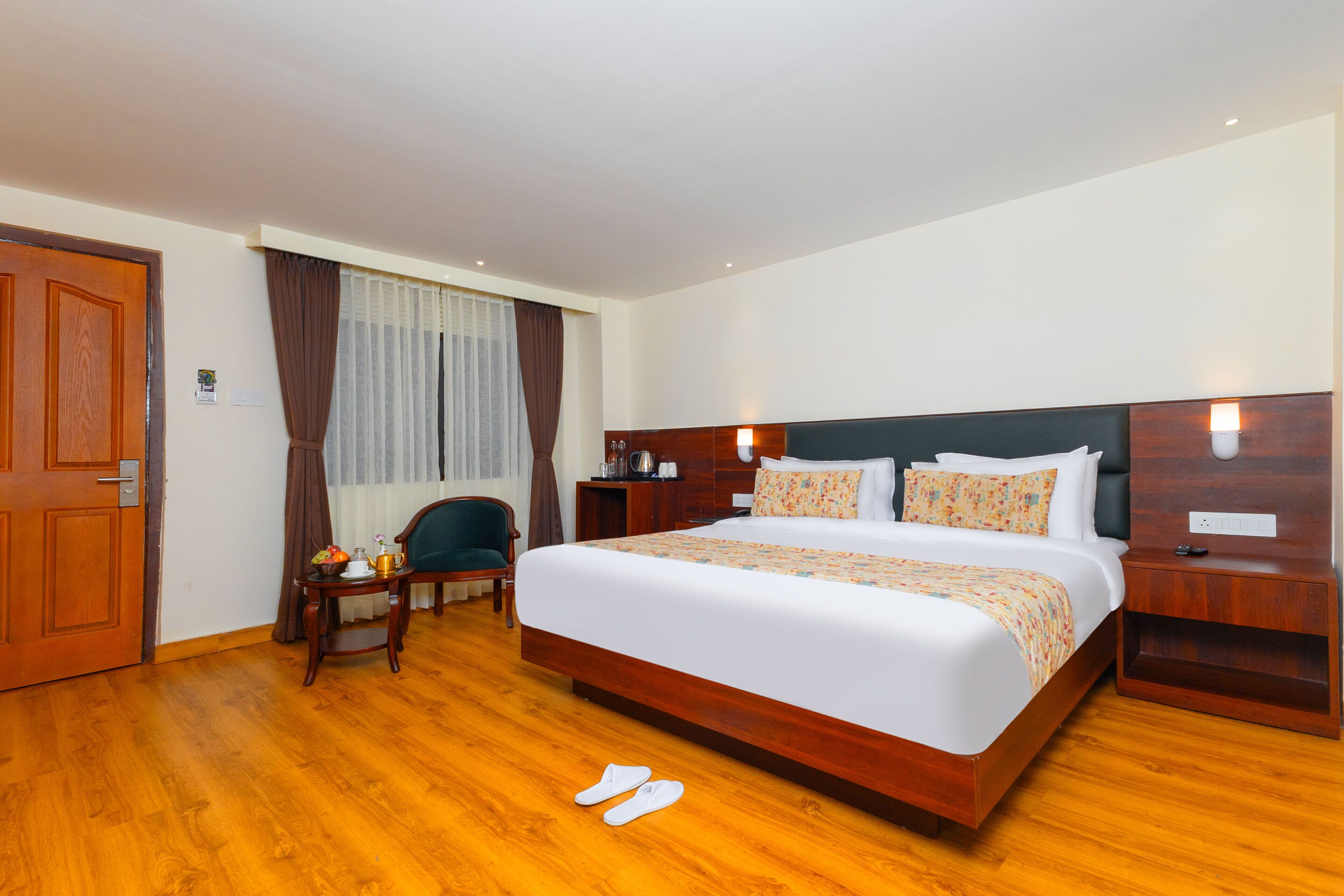 A hotel room with a wooden floor, a large bed with a headboard, a bedside table, a chair, and a door.
