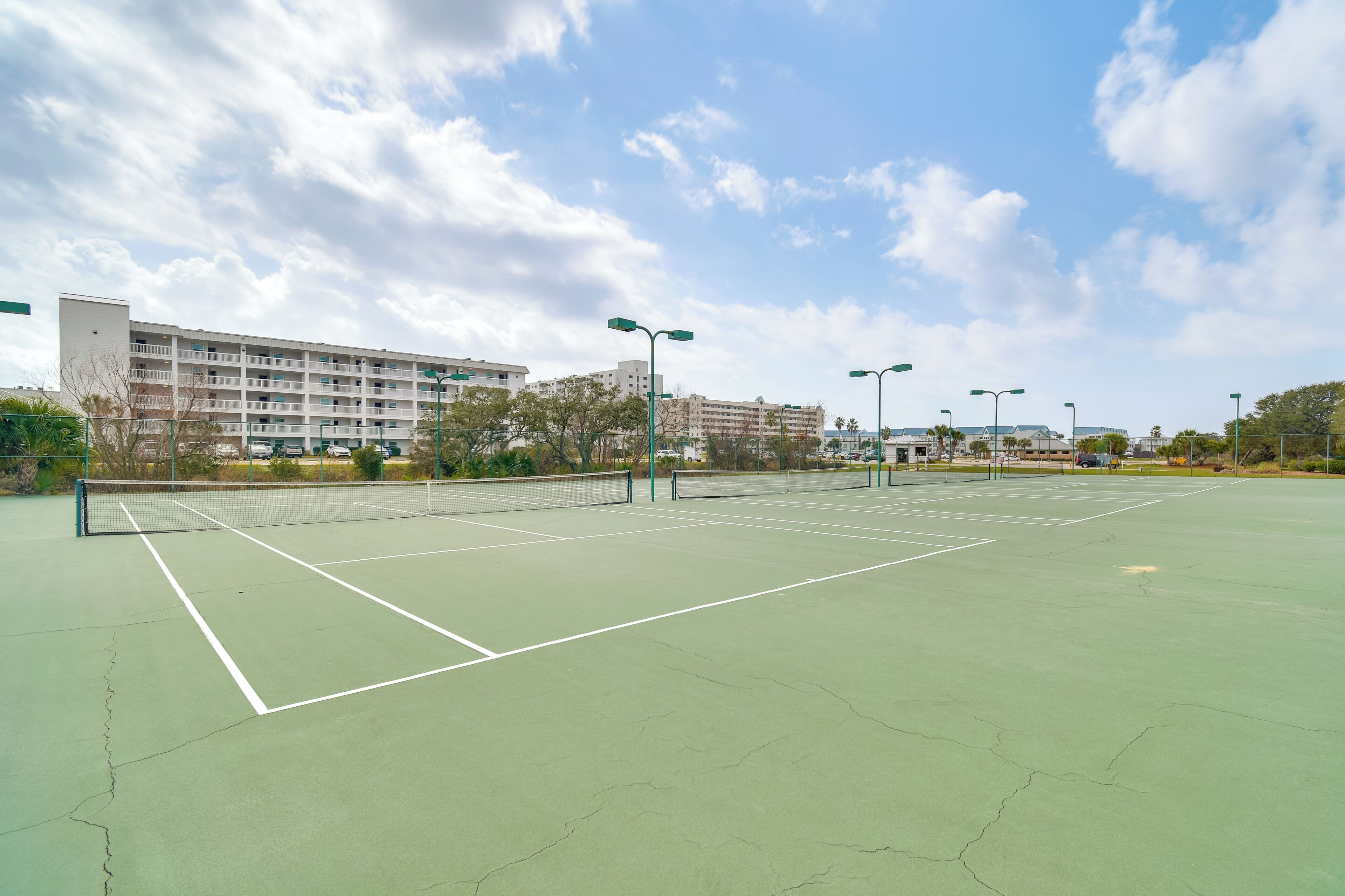 Una cancha de tenis al aire libre con una red, rodeada por una valla, y edificios al fondo.